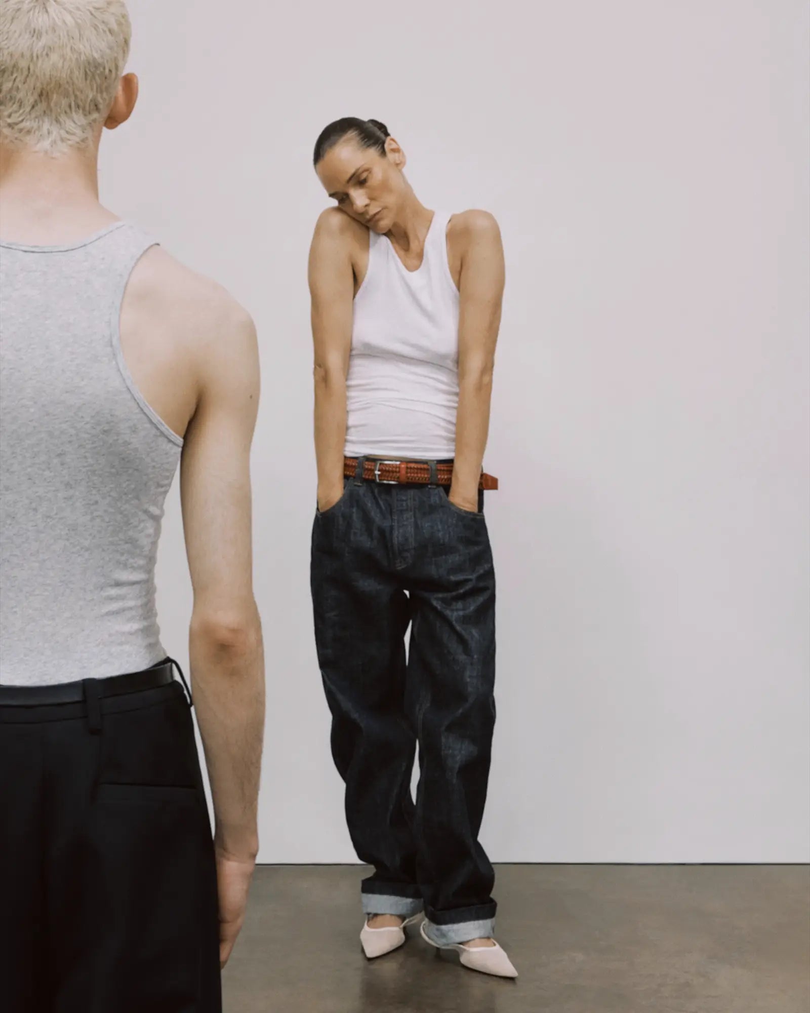 Two people standing side by side, one wearing a gray tank top and black pants, the other in a white tank top and dark jeans.